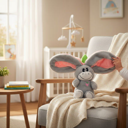 Momviacare grey plush toy with large floppy pink-lined ears, embroidered face, and strawberry detail, sitting on a chair with a blanket.